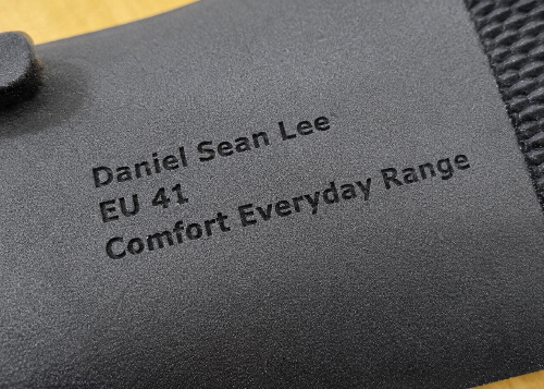 Close-up of a dark gray fabric strap or band with black printed text reading 'Daniel Sean Lee', 'EU 41', and 'Comfort Everyday Range', next to a textured knit section.