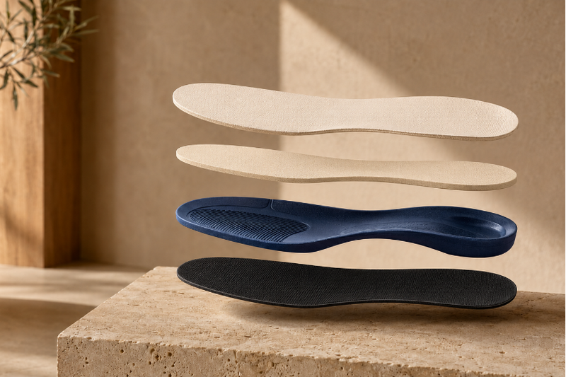 Three orthopedic shoe insoles stacked vertically in different colors: cream, navy blue, and black, floating above a stone pedestal in a warm, minimalist room setting.